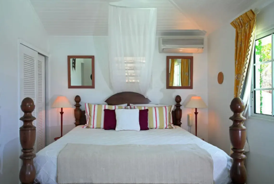 A dark wood four-poster bed with sheer white canopy anchors this serene bedroom. Striped and maroon pillows rest on crisp white linens, while two wooden-framed mirrors hang above. Yellow curtains and garden views through the window add warmth—classic Caribbean elegance.