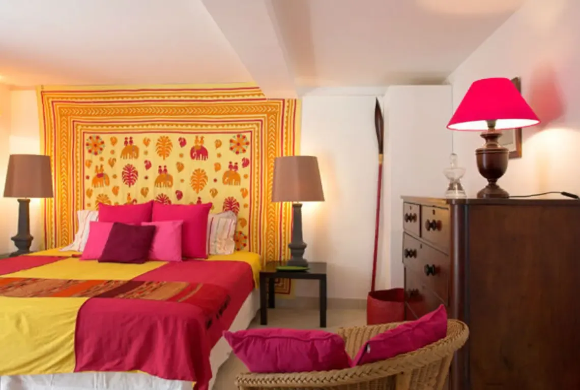 Bright yellow and magenta linens dress the bed beneath a colorful tapestry with elephant motifs and geometric patterns. A wicker chair with magenta cushions and a dark wooden dresser with red-shaded lamp add eclectic warmth—this bedroom is a vibrant, globally-inspired retreat.