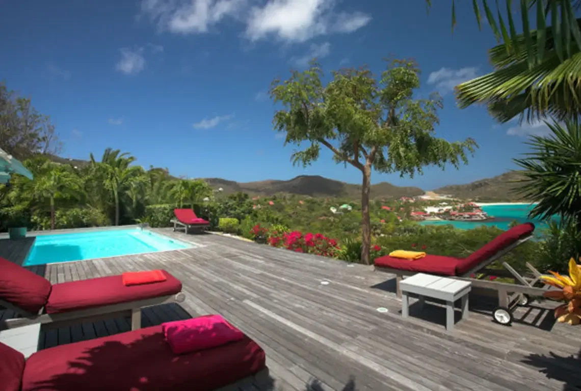 Red lounge chairs with colorful cushions line a spacious wooden deck beside a brilliant blue pool. Lush palms and blooming flowers frame the scene, while rolling hills and a turquoise ocean stretch to the horizon—the ultimate outdoor living room.