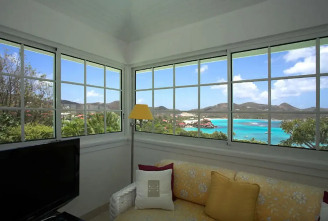 Floor-to-ceiling windows frame a breathtaking turquoise ocean panorama in this light-filled living space. A yellow floral sofa with colorful cushions invites you to linger while the Caribbean unfolds before you—nature's own masterpiece as your living room wallpaper.