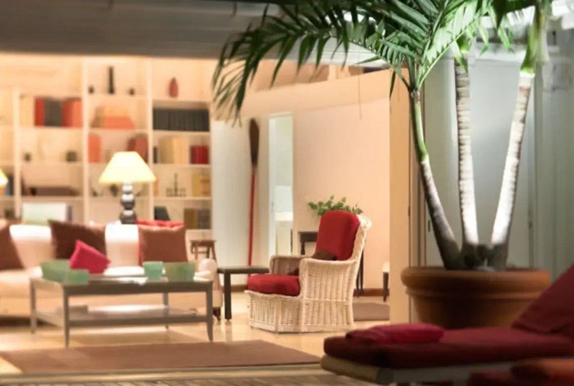 A potted palm and wicker armchair with red cushion anchor the corner of this inviting living room. A light sofa with colorful pillows sits before a built-in bookshelf filled with books and treasures, while a warm lamp glow creates the perfect evening retreat.