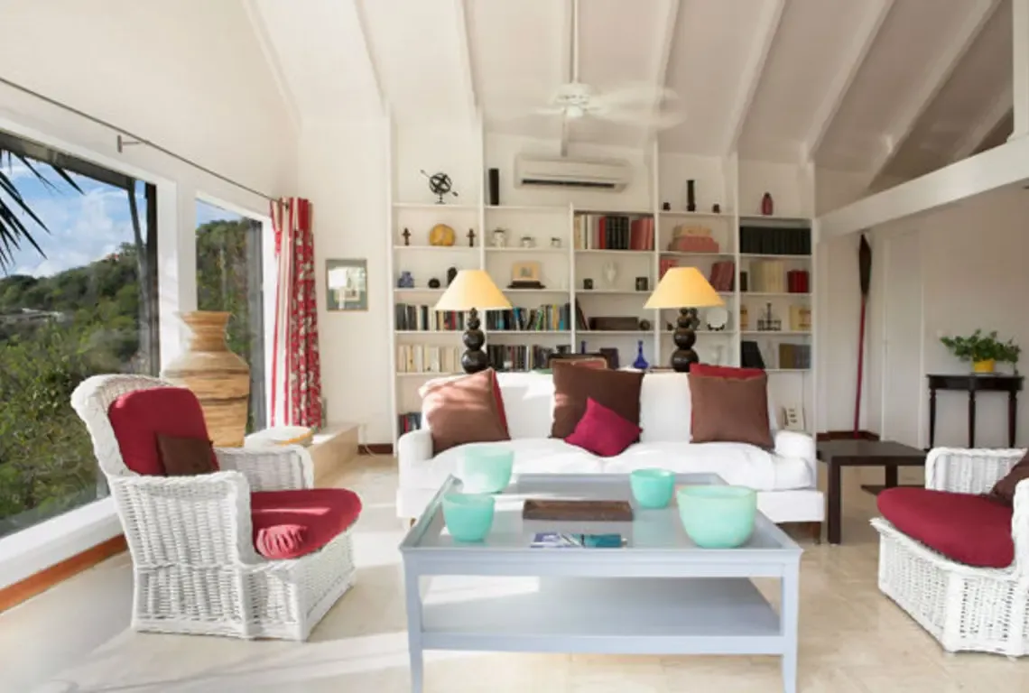 High white-beamed ceilings and a ceiling fan crown this airy living room where a white sofa with red pillows faces a light blue coffee table with turquoise bowls. White wicker armchairs and a built-in bookshelf add warmth while large windows frame lush tropical greenery.