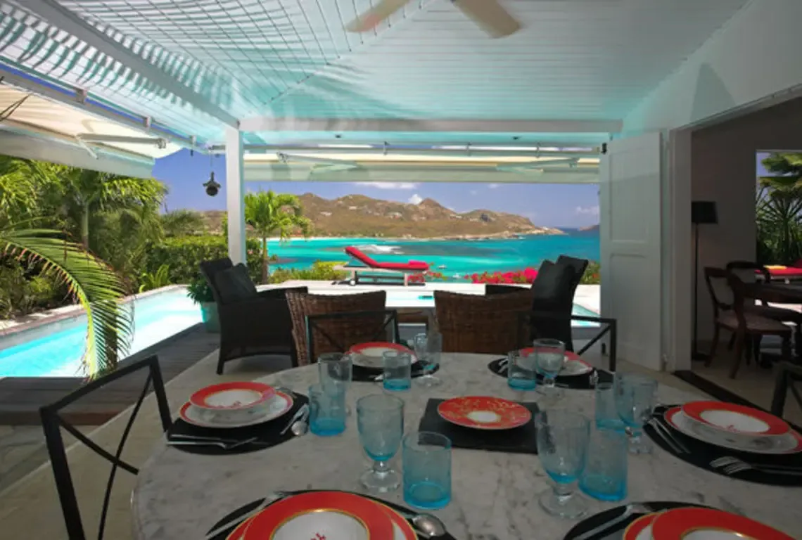 A round marble table set with red-and-white plates and blue glasses sits under a translucent canopy, while the turquoise ocean and hilly islands stretch beyond. Pink flowers and palm fronds frame this open-air dining experience—every meal comes with a million-dollar view.