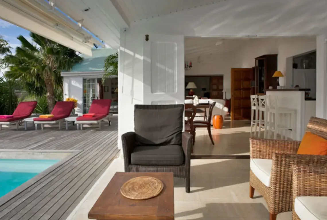 Red lounge chairs with colorful towels line the pool deck, while a covered patio with wicker armchairs bridges the gap between turquoise pool and bright open-concept interior. White walls, warm wood, and filtered sunlight create effortless indoor-outdoor Caribbean living.