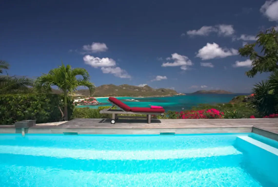 A turquoise pool with shallow steps mirrors the Caribbean sky, while a red chaise lounge on the wooden deck faces an endless ocean panorama. Pink bougainvillea and palm trees frame the scene—pure tropical luxury where every angle is a postcard.