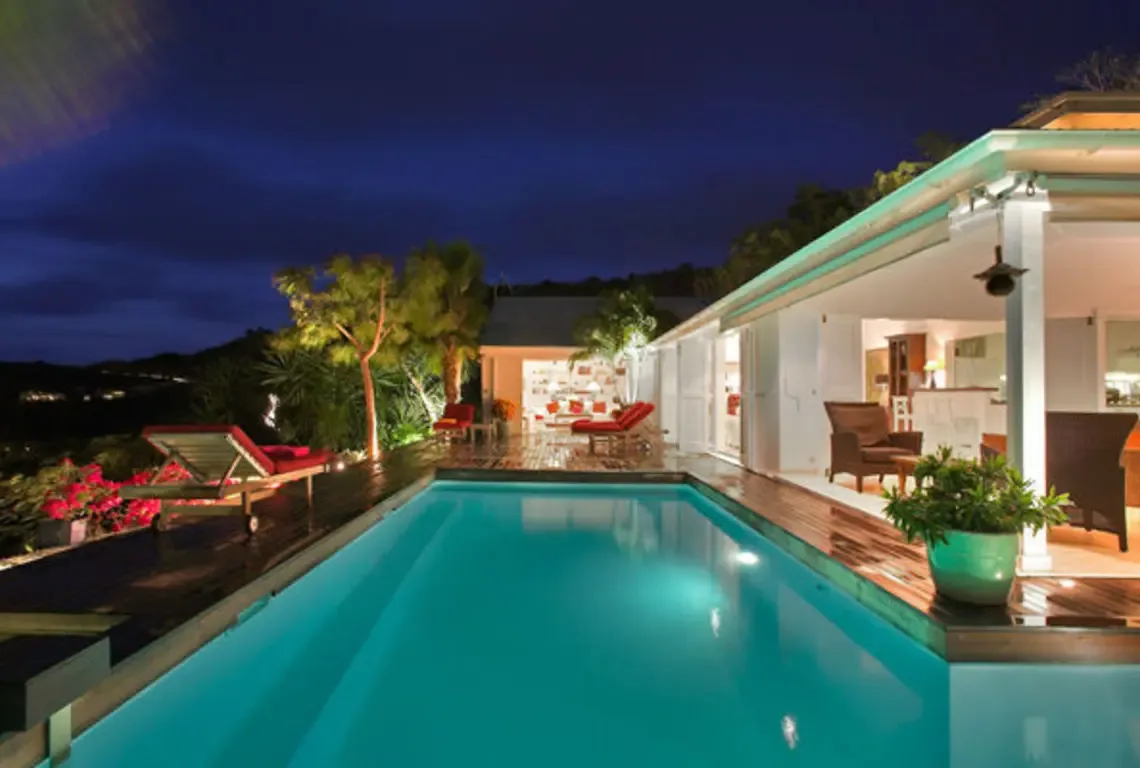 The illuminated turquoise pool glows under the night sky as warm interior light spills through sliding glass doors. Red lounge chairs and potted plants line the wooden deck while distant hillside lights twinkle—Caribbean evenings don't get more magical than this.