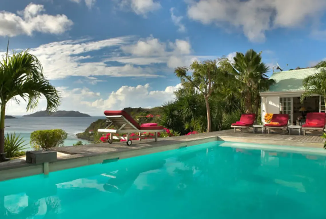 A turquoise swimming pool reflects the Caribbean sky while red lounge chairs line the wooden deck. Beyond, the calm ocean stretches to distant islands under fluffy clouds—your private paradise where every day feels like a painting.
