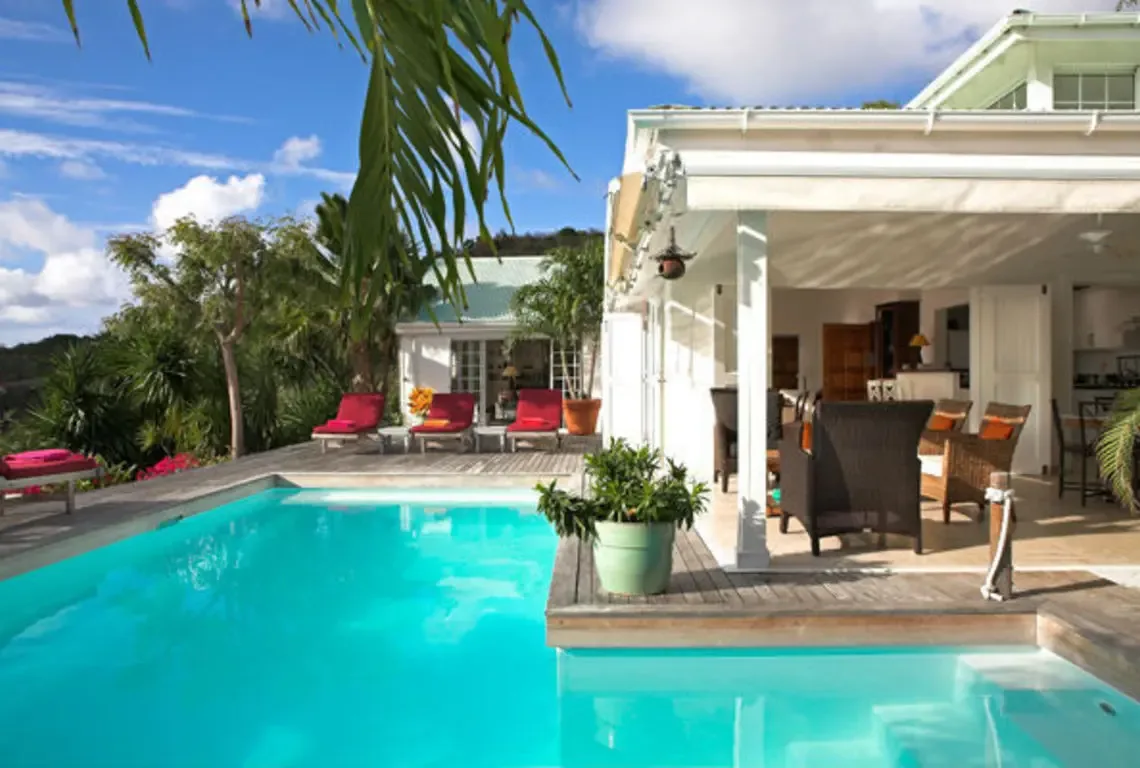 Red lounge chairs line the wooden deck beside a brilliant turquoise pool, while a covered patio with wicker furniture invites outdoor dining. Pink blooms and palm trees frame the white villa with green roof—indoor-outdoor living at its Caribbean finest.