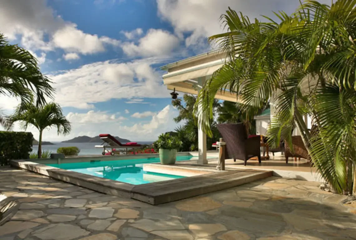 A turquoise pool reflects vivid blue sky on a stone-tiled deck with a red-cushioned lounger facing the ocean and distant islands. A covered veranda with wicker armchair invites shaded relaxation while palms sway overhead—every detail designed for effortless island living.