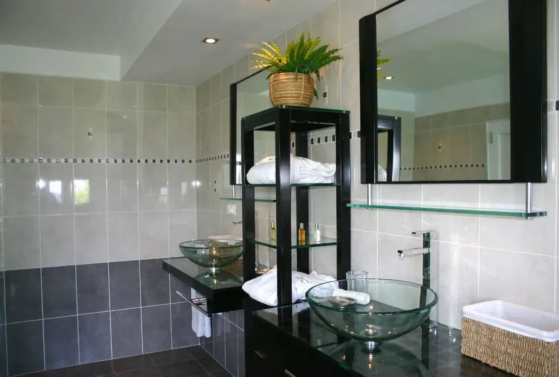 Two glass vessel sinks gleam atop a dark glossy countertop with modern chrome faucets. A black-framed shelving unit with crisp white towels and a wicker basket topped with greenery adds warmth, while twin mirrors and recessed lighting complete this sleek spa-inspired bathroom.