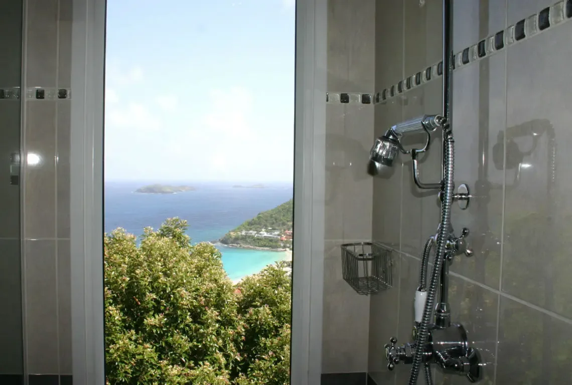 A sleek chrome shower with glass panels and mosaic tile detail opens to an unobstructed view of turquoise waters and a crescent beach. Lush green foliage cascades down the hillside while distant islands dot the horizon—the most scenic shower you'll ever take.
