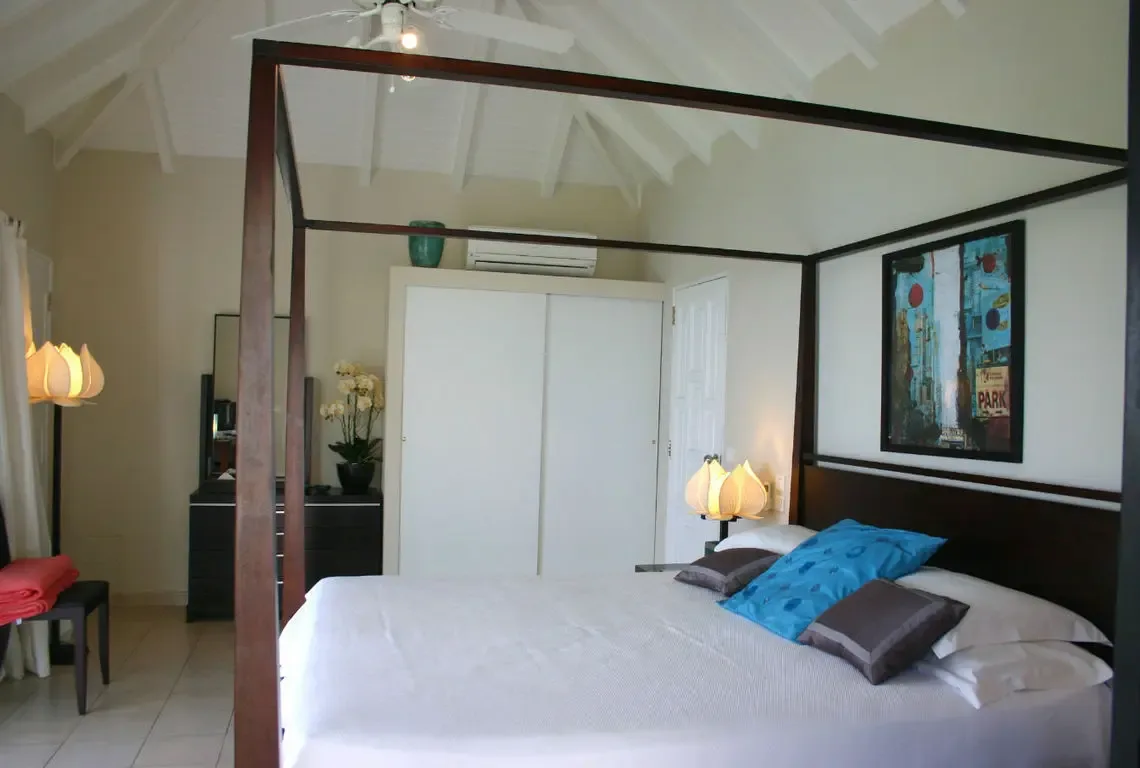 A dark wood four-poster bed with white linens and a blue accent pillow anchors this serene bedroom beneath vaulted white-beamed ceilings. A colorful abstract artwork hangs above while a white orchid on the dresser and flower-shaped lamps add refined elegance—peaceful Caribbean nights await.