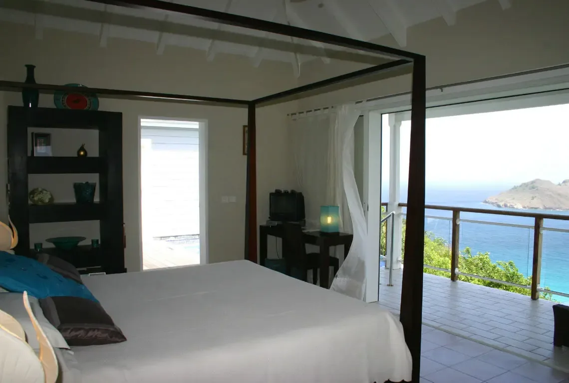 A dark wood four-poster bed with white linens and blue accent pillows sits beneath vaulted white beams. Sliding glass doors open to a balcony overlooking the ocean and rocky island, while a curated shelf with blue vases and golden bowls adds personality—wake up to the Caribbean every morning.