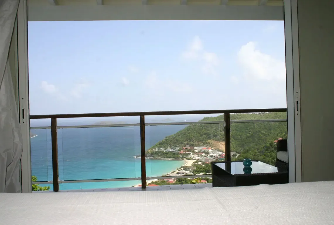 Crisp white linens dress a bed that faces sliding glass doors opening to a jaw-dropping ocean panorama. A dark wicker chair with turquoise vase sits beside the view where turquoise shallows meet a crescent beach backed by lush green hills—this is what waking up in paradise looks like.