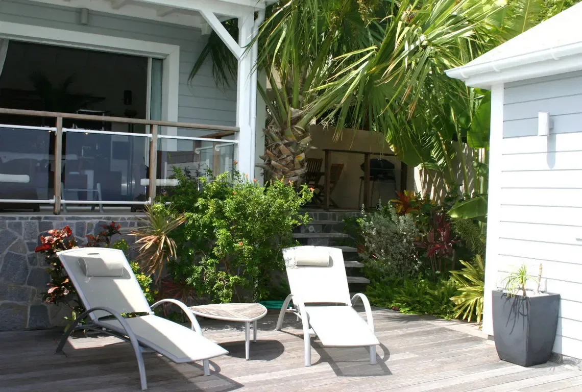 Two white lounge chairs with a round side table sit on a weathered wooden deck beside the villa's glass-walled interior. Vibrant red and green tropical foliage wraps around the stone walls—your private garden retreat where afternoon naps come naturally.