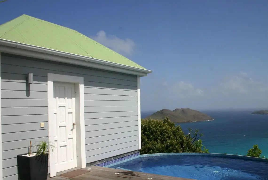 Light grey siding and a pale green roof define this hillside villa where an infinity pool with blue mosaic tiles merges with the Caribbean. Lush islands rise from turquoise shallows while white trim and a wooden deck complete this picture-perfect retreat above the sea.