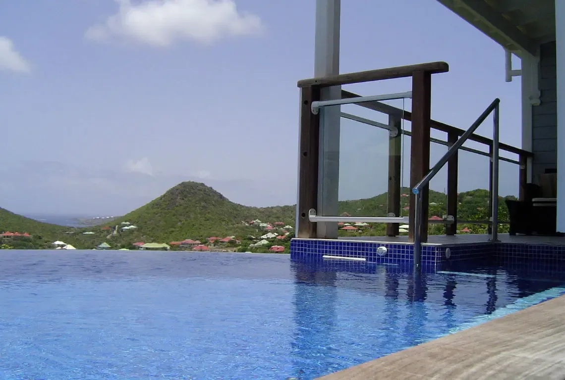 A wide infinity pool with dark blue tile edge merges with the horizon, while a modern glass railing and wooden deck frame the foreground. Colorful houses with red and green roofs dot the lush hillside beyond—your private perch above Caribbean paradise.