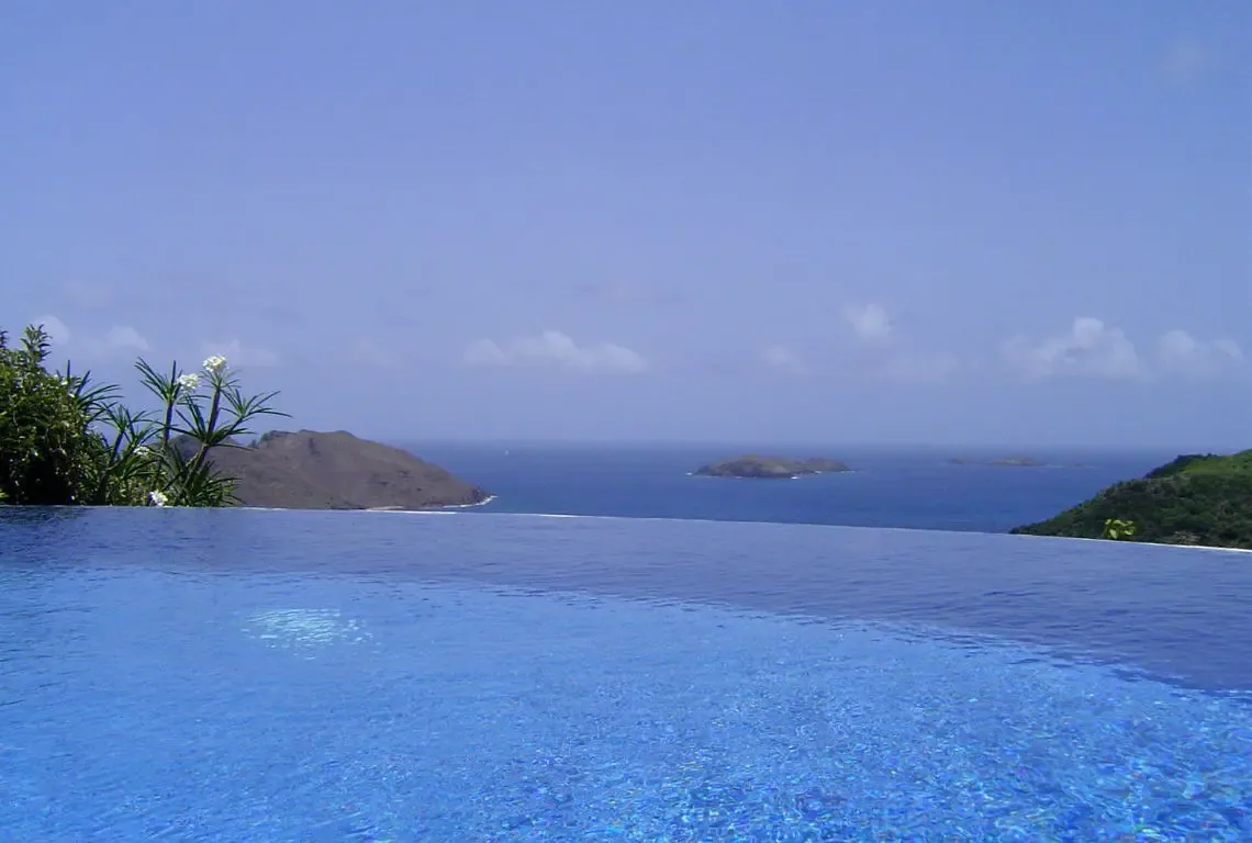 The blue infinity pool merges seamlessly with the ocean where three small islands dot the deep blue sea. White plumeria blossoms frame the left while a lush green coastline curves to the right—where the pool ends and the Caribbean begins is impossible to tell.