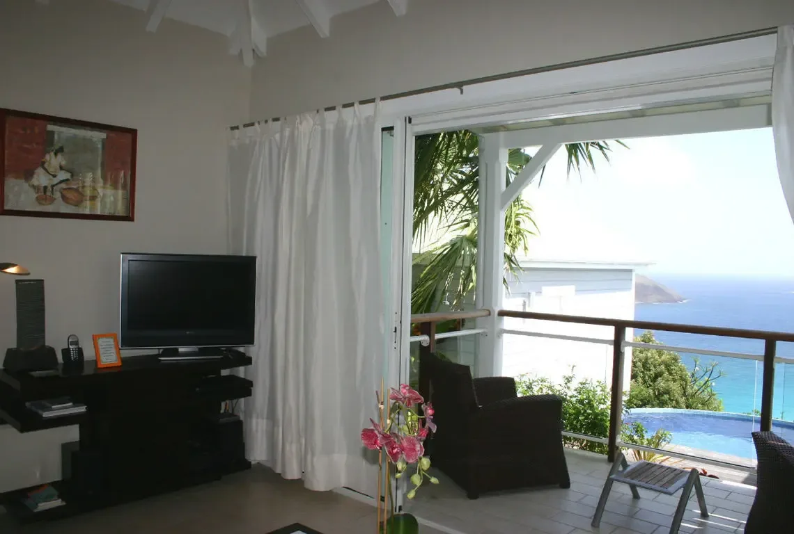 White curtains frame sliding glass doors that open to a balcony with wicker chairs overlooking the pool and ocean. Pink orchids in a tall vase bring tropical color indoors while a flat-screen TV and framed art complete this living room where the Caribbean is always in frame.