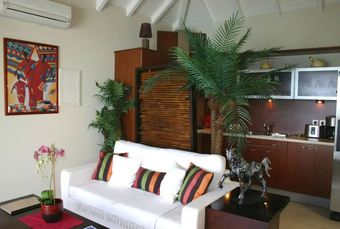 A white sofa with vibrant striped pillows faces a dark wood coffee table topped with a potted pink orchid. A metallic horse statue adds artistic flair beside tropical plants, while a colorful geometric painting and vaulted white beams complete this stylish Caribbean living space.