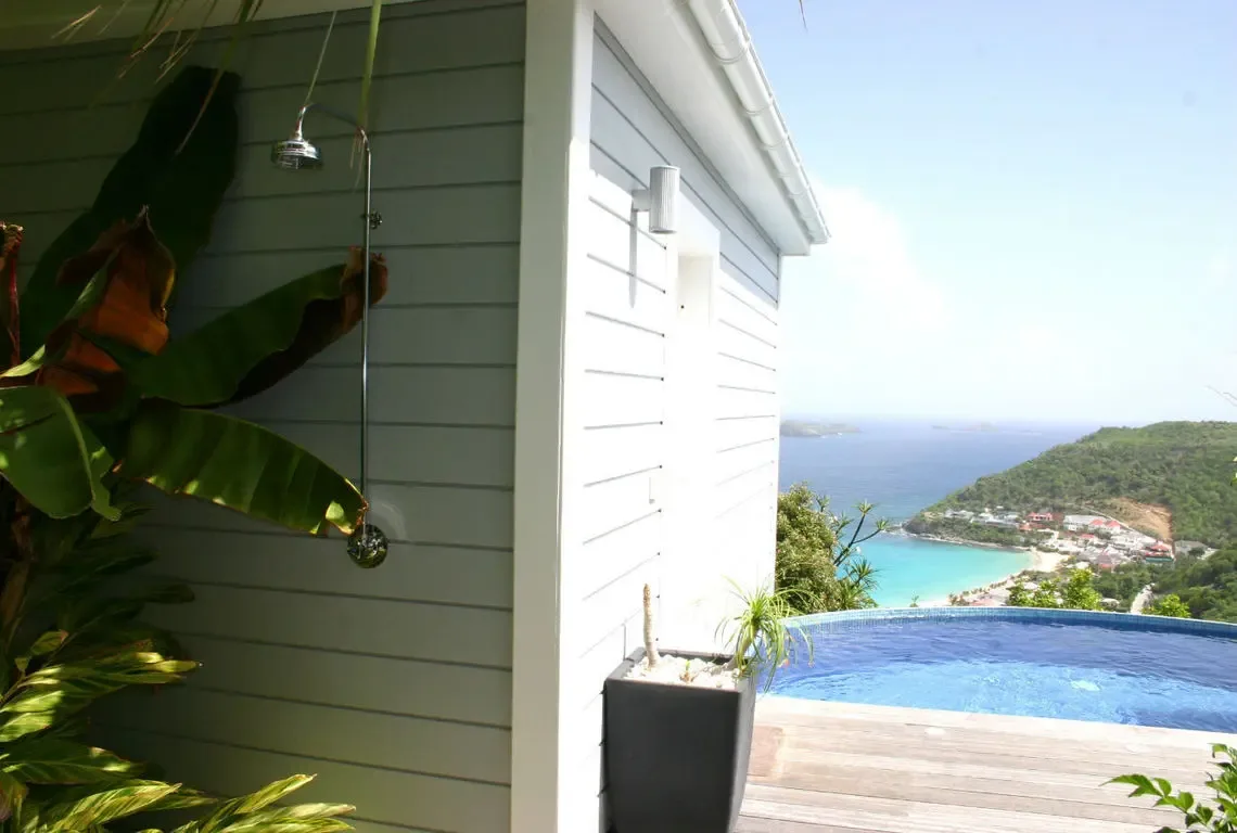 An outdoor shower framed by lush banana leaves sits beside the wooden deck leading to the infinity pool. Beyond, a turquoise bay curves around a sandy beach with a cluster of seaside buildings—rinse off after a swim with the Caribbean as your backdrop.