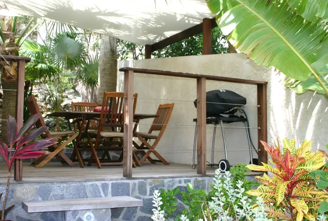 A wooden dining table with slatted chairs sits on a stone-tiled patio beneath a light canopy, while a black gas grill stands ready for Caribbean cookouts. Lush crotons in red, yellow, and green frame the scene with palm fronds overhead—alfresco dining surrounded by tropical color.