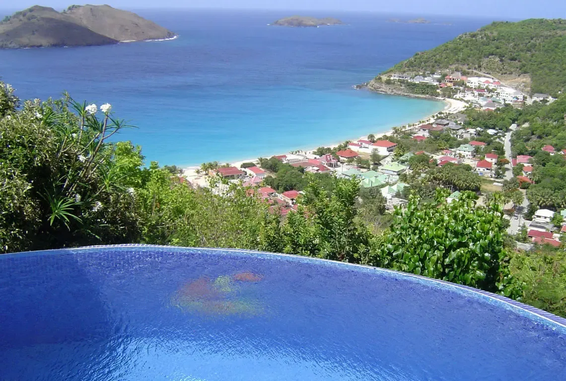A curved blue infinity pool overlooks a crescent beach and charming coastal town with red and green roofs. Turquoise shallows fade to deep blue ocean where distant islands dot the horizon—Colombier Bay unfolds beneath you like a living painting.