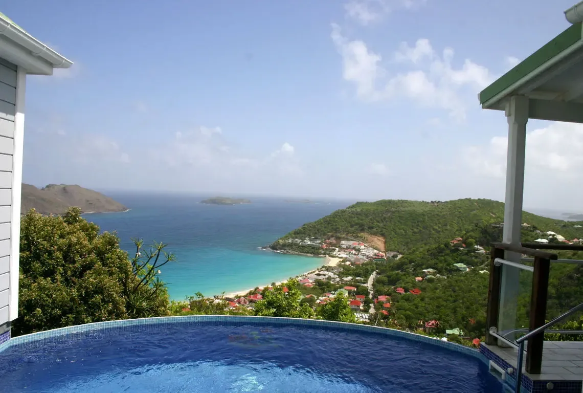 A circular blue-tiled pool sits at the edge of the terrace, overlooking a crescent beach where turquoise shallows meet deep blue sea. A village with red-tiled roofs nestles in the green valley while distant islands rise from the horizon—this is the view you'll never forget.