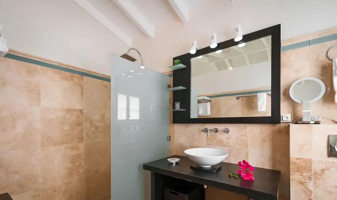 A dark wood vanity with a white vessel sink sits beneath a black-framed mirror on tan stone walls, where a pink bougainvillea flower and a frosted glass rain shower accent the spa-like space.