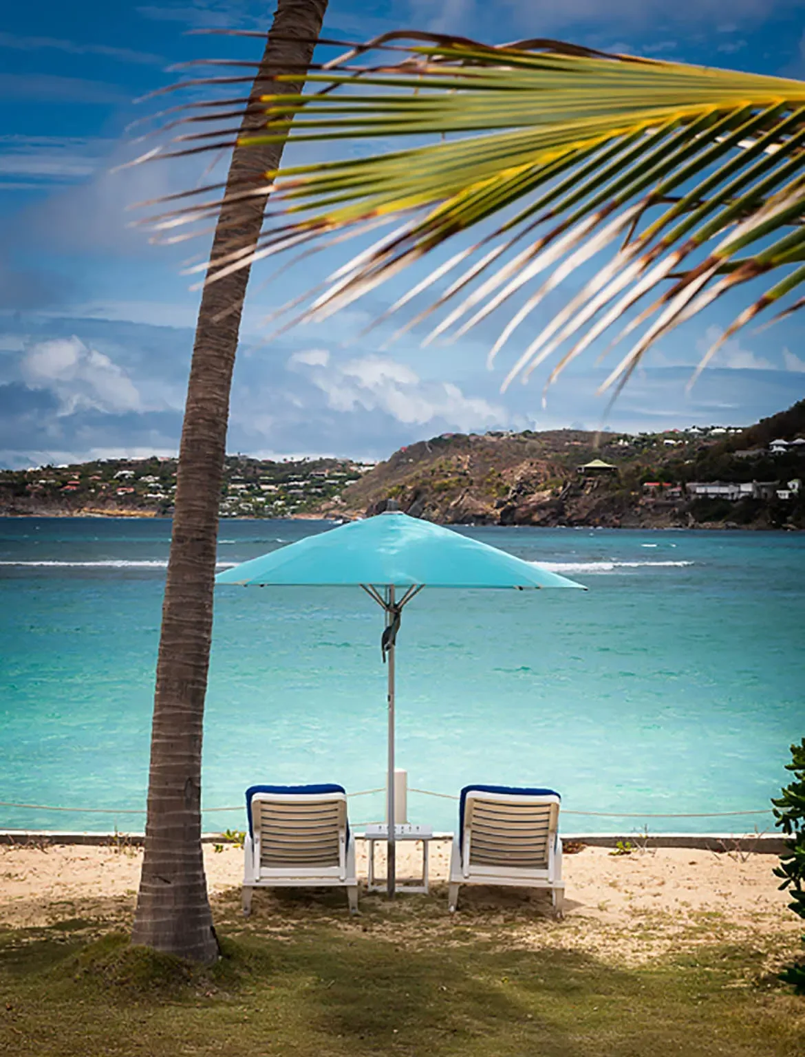 Two white lounge chairs with blue cushions sit beneath a turquoise umbrella beside a palm tree, where crystalline turquoise water meets a white sand beach and sun-drenched hills dot the horizon.
