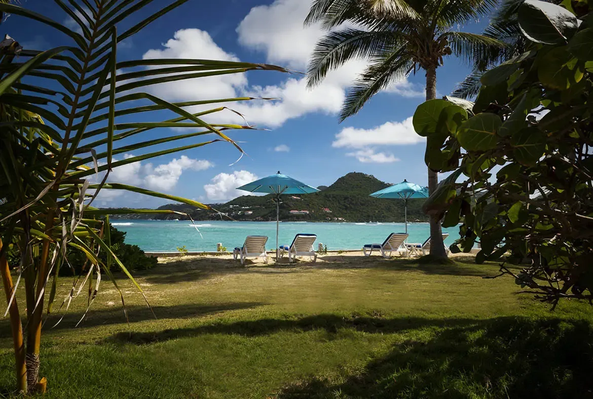 White lounge chairs beneath turquoise umbrellas sit on a green lawn, where palm fronds frame a crystalline turquoise bay with white villas dotting lush green hills beneath puffy clouds.