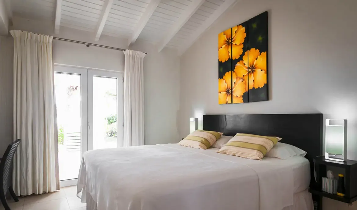 A white bed with striped pillows sits beneath a vibrant yellow hibiscus triptych on a vaulted-white-beam ceiling wall, where French doors with white curtains open to lush tropical greenery.