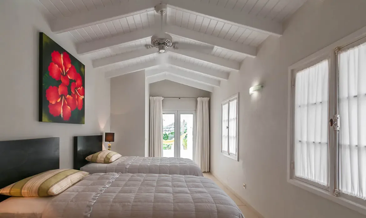 Two twin beds with grey quilts sit side by side beneath a vaulted white-beam ceiling, where a vibrant red hibiscus painting and French doors opening to a sunlit garden accent the bright room.