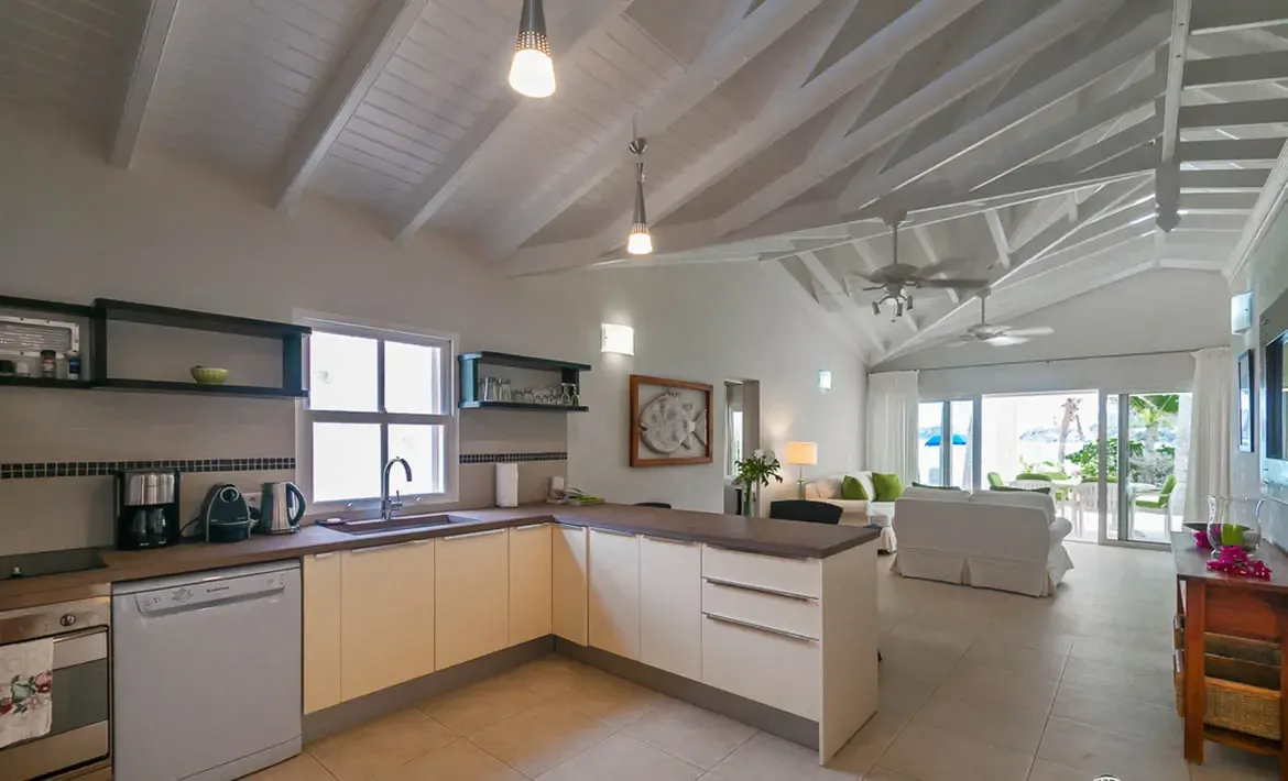 A modern kitchen with cream cabinets and dark countertops sits beneath a vaulted white-beam ceiling, where teardrop pendant lights and glass doors open to a covered patio surrounded by lush tropical greenery.
