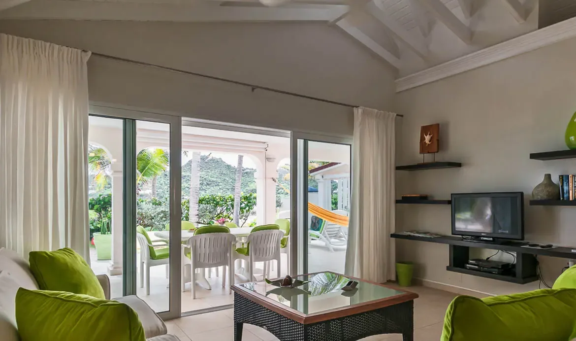 A white sofa with lime-green pillows sits beneath a vaulted white-beam ceiling, where open glass doors lead to a covered patio with a dining table and an orange hammock amid tropical gardens.