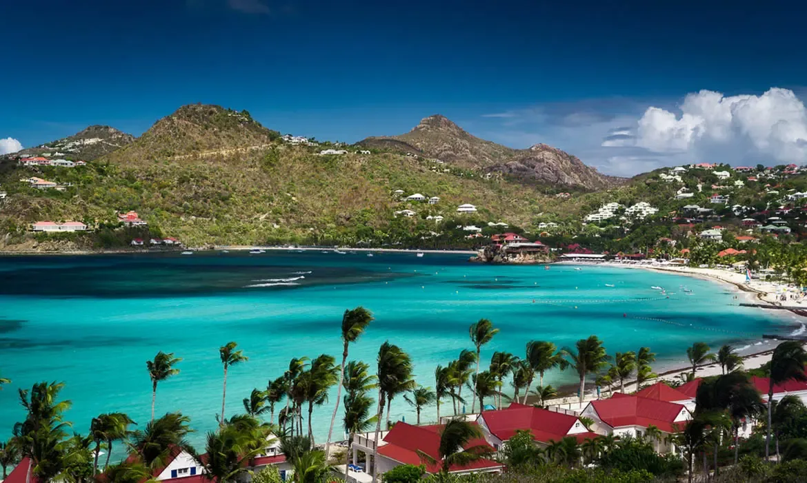 Red-roofed villas nestle among palms above turquoise Saint Jean Bay, where Eden Rock perches on a rocky outcrop and green hills rise across the crescent-shaped coast.