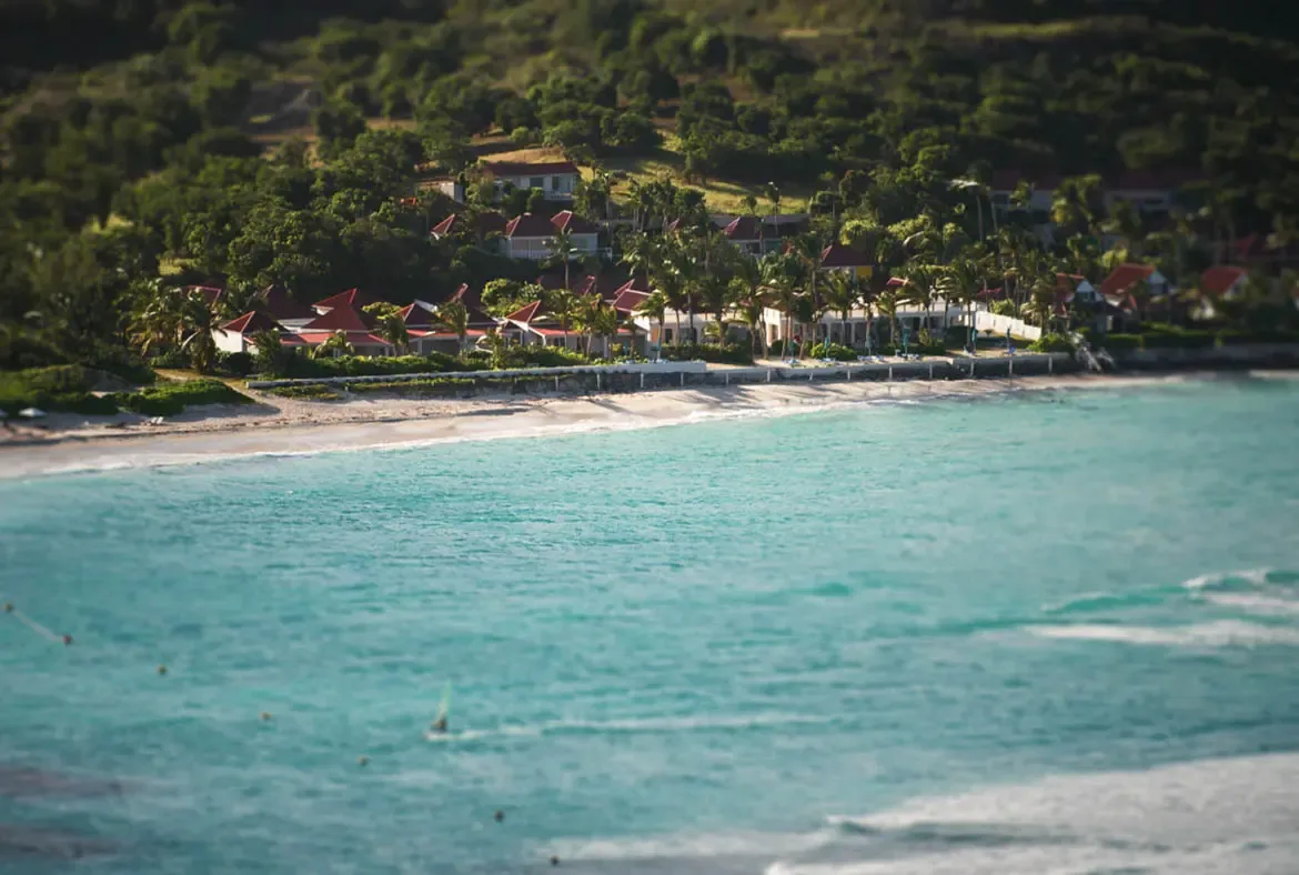 An aerial tilt-shift view captures red-roofed villas nestled between a lush hillside and turquoise ocean, where a narrow sandy beach meets crystal-clear Caribbean water.