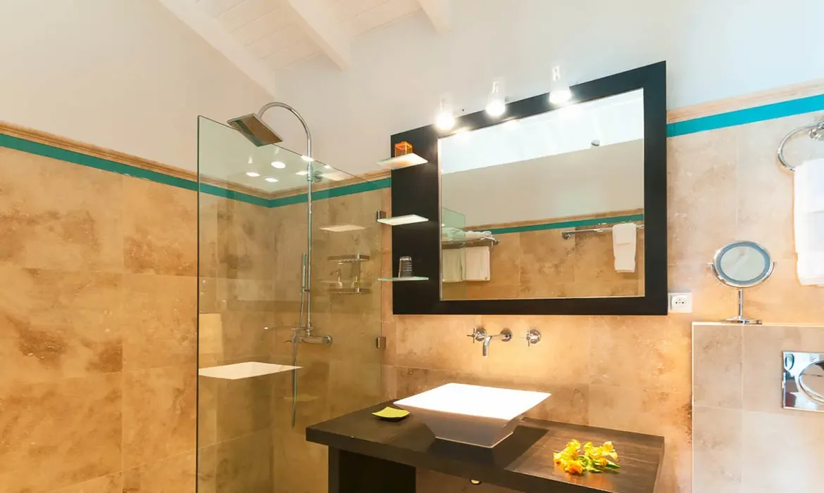 A dark wood vanity with a rectangular vessel sink sits under a framed mirror and globe lights, where a glass-enclosed rainfall shower and travertine walls with a turquoise accent stripe create a spa-like retreat.