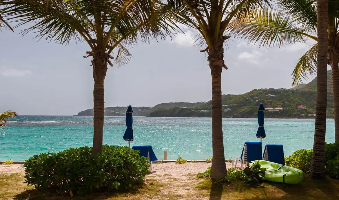 Blue beach umbrellas and loungers rest under palms beside a lime-green kayak on grass, where turquoise water and green hills dotted with villas stretch across a Caribbean bay.