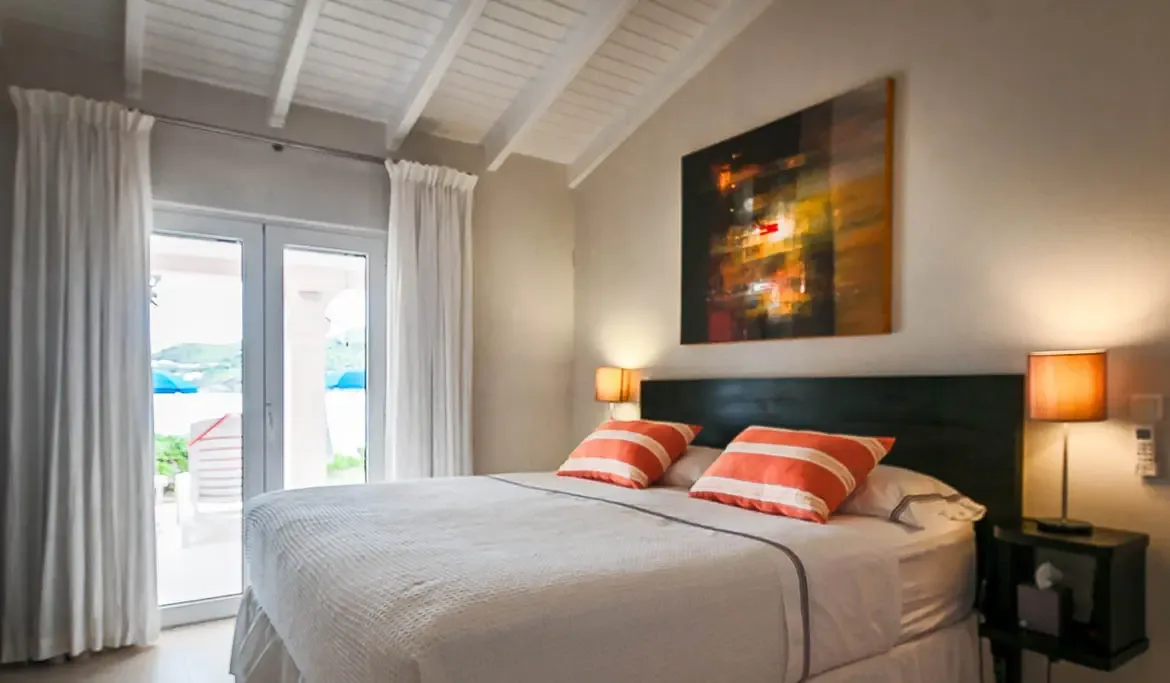 A white bed with orange-striped pillows and a warm abstract painting sits under a vaulted ceiling, where French doors open to a sunlit terrace with glimpses of blue.
