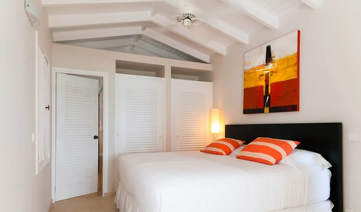A white bed with orange-striped pillows and a fiery red abstract painting sits under a vaulted ceiling, where white louvered closet doors and a tall lamp add clean modern comfort.