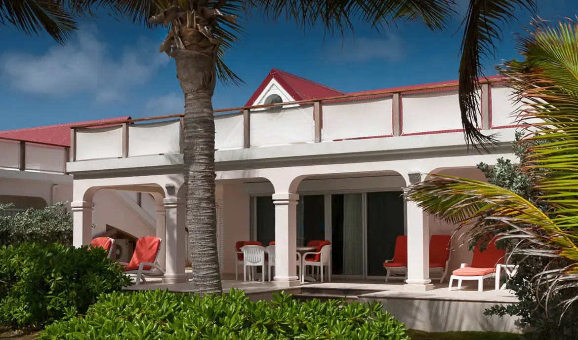 A two-story white villa with a red gabled roof and arched white columns shelters orange-cushioned seating, where palm trees frame the tropical facade under a blue sky.