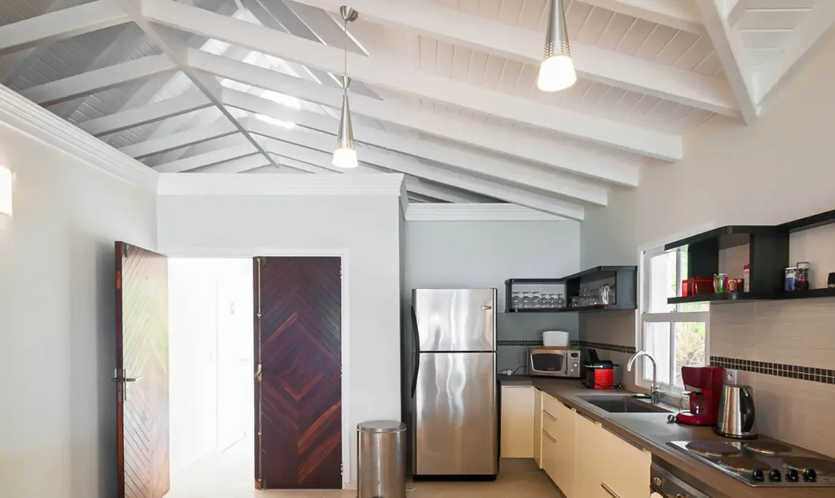 A modern kitchen with cream cabinetry and dark countertops sits under a white vaulted ceiling, where chevron-patterned doors open to sunlight and a red coffee maker adds a pop of color.