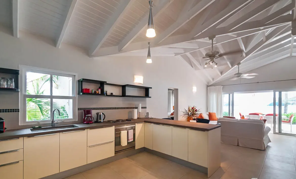A modern kitchen with cream cabinetry and dark countertops sits under a white vaulted ceiling with pendant lights, where a red coffee maker and open shelves add warmth near a window looking onto tropical greenery.