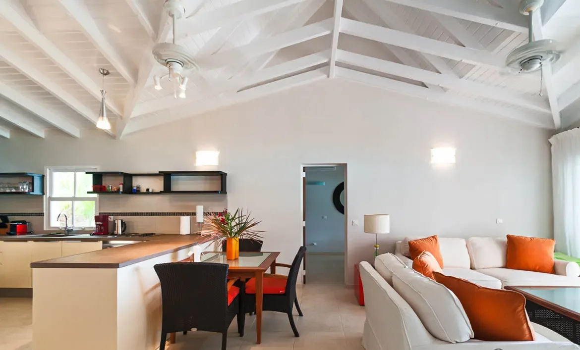 Cream sofas with orange pillows and a wicker coffee table sit under a vaulted ceiling, where a dining table with orange-seat chairs connects to a modern kitchen in an open-concept layout.