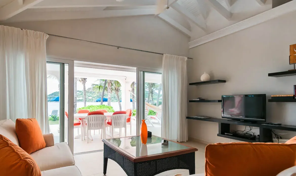 Cream sofas with orange pillows and a glass coffee table sit under a vaulted ceiling, where sheer curtains frame a covered terrace and turquoise ocean beyond.