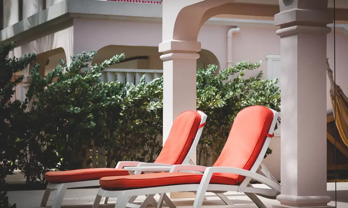 White loungers with orange cushions sit under arched columns beside a striped hammock, where pink walls and lush hedges frame a garden view.