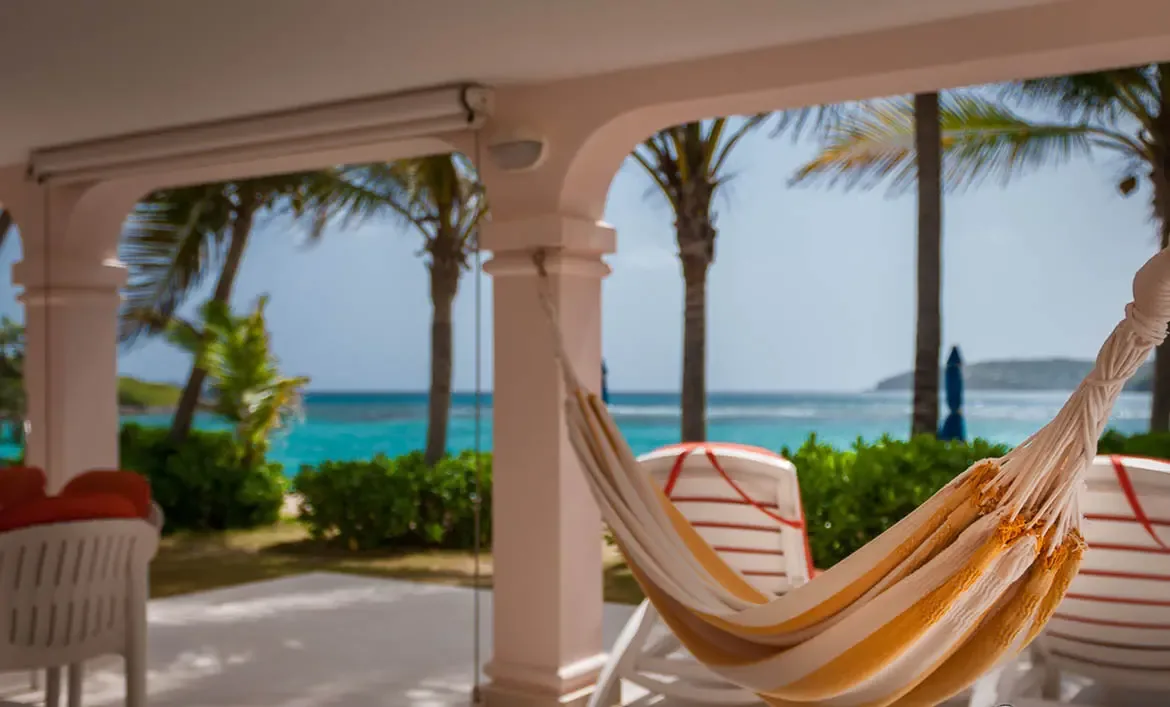 An orange-striped hammock hangs from a shaded porch with white arches, where palm trees and turquoise ocean create a breezy indoor-outdoor retreat.