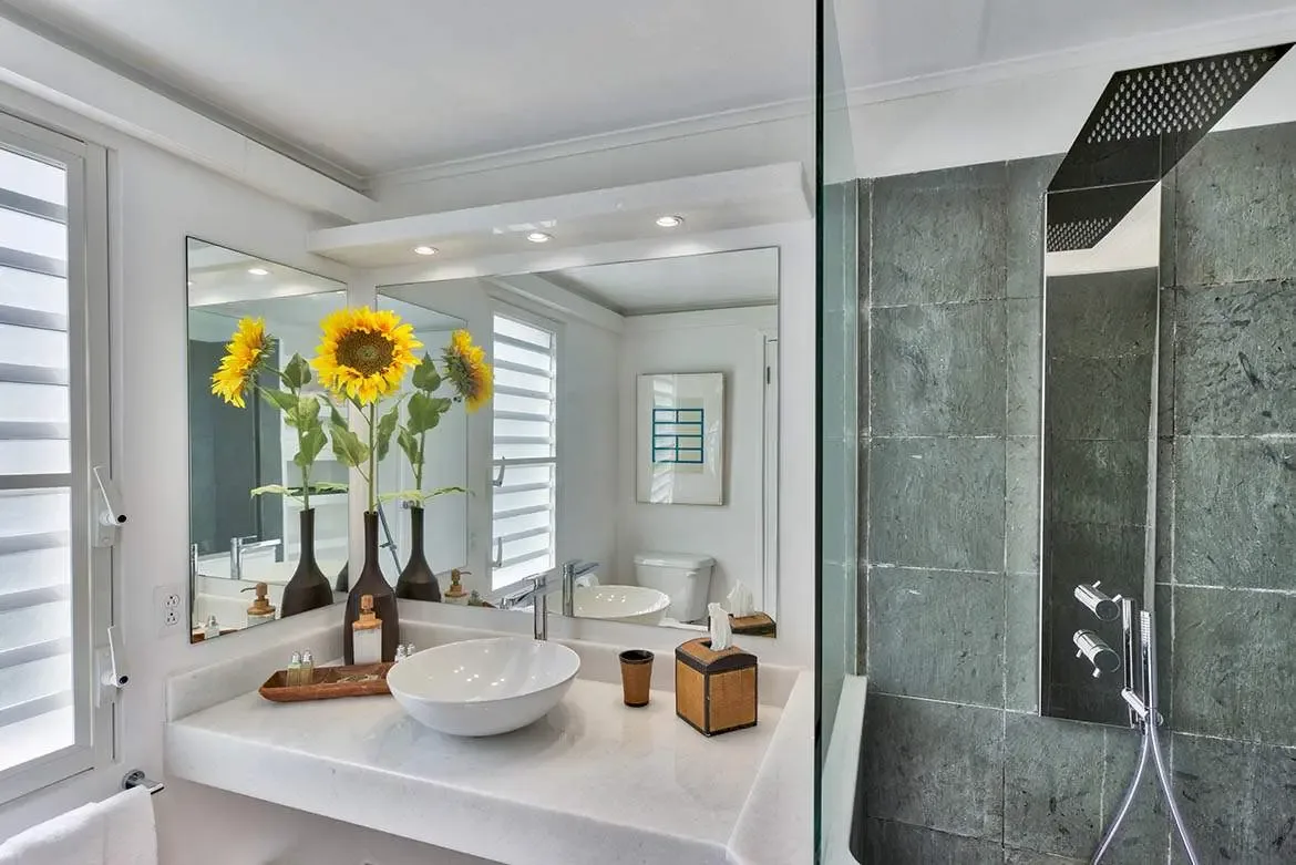 A white floating vanity with a vessel sink holds three yellow sunflowers in brown vases beside a frameless mirror, where a glass-panelled shower with dark slate walls and a rain showerhead adjoins.