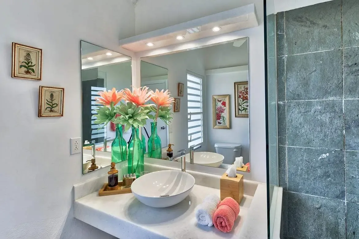 A white marble vanity with a vessel sink holds coral flowers in green glass bottles beside a frameless mirror, where a glass-panelled shower with grey slate tiles and a rain showerhead adjoins the bright space.